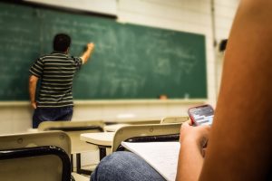Teacher writing on a blackboard while you see a student texting in the forefront.